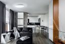 Modernes Wohnzimmer mit schwarzem Sofa, offener Küche, Holzboden und großen Fenstern, stilvoll eingerichtet.