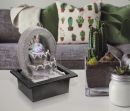 Moderner Zimmerbrunnen mit LED-Kugel auf weißem Tisch, daneben Kaktus, Kerze und Sofa mit Pflanzenkissen.