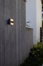 Moderne Außenwandleuchte an grauer Betonwand, beleuchtet, mit Gartenpflanzen im Vordergrund, minimalistisches Design.
