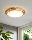 Moderne LED-Deckenleuchte mit goldener Verzierung leuchtet in hellem Wohnzimmer mit Fenster und Pflanze.